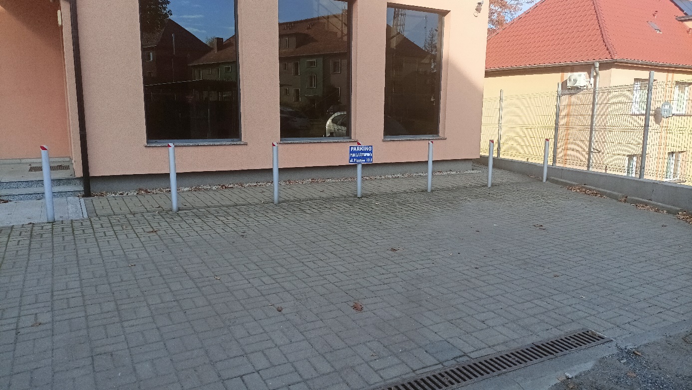 Ilustracja przedstawia parking przed budynkiem organu. Przed budynkiem na metalowym słupku tabliczka z napisem ,,Parking dla użytkowników Piastów 10J
